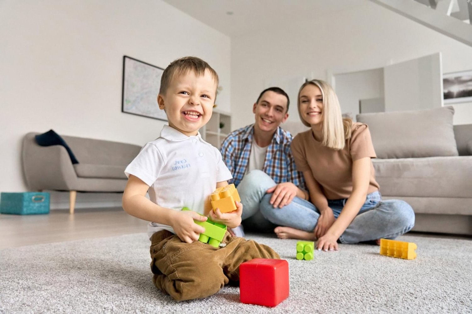 Toddler smiling with plastic blocks while parents watch in a bright living room